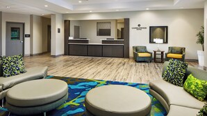 Zona de recepción de un hotel con mostrador, área de asientos con sillas y otomanas, y alfombra estampada.