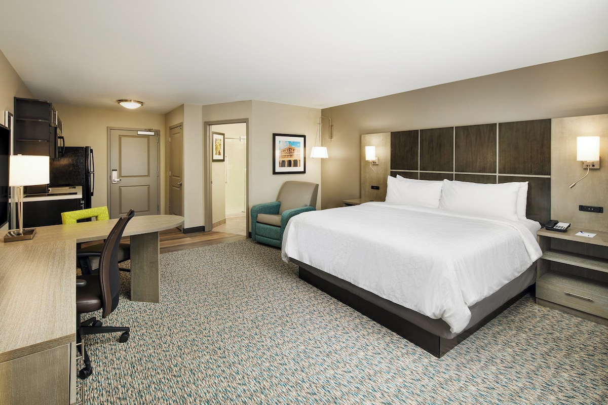 A hotel room with a large bed, a desk with a chair, a small armchair, a desk lamp, and a nightstand with a phone.