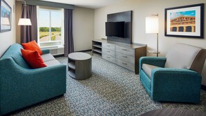 Una habitación de hotel moderna con un sofá color turquesa, un sillón más pequeño, un televisor de pantalla plana, una mesa redonda pequeña y un ventanal con cortinas.