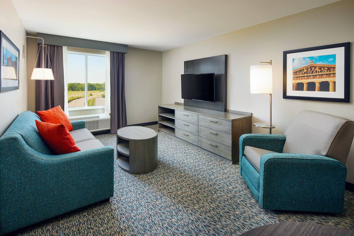 A modern hotel room with a teal sofa, a smaller armchair, a flat-screen TV, a small round table, and a large window with curtains.