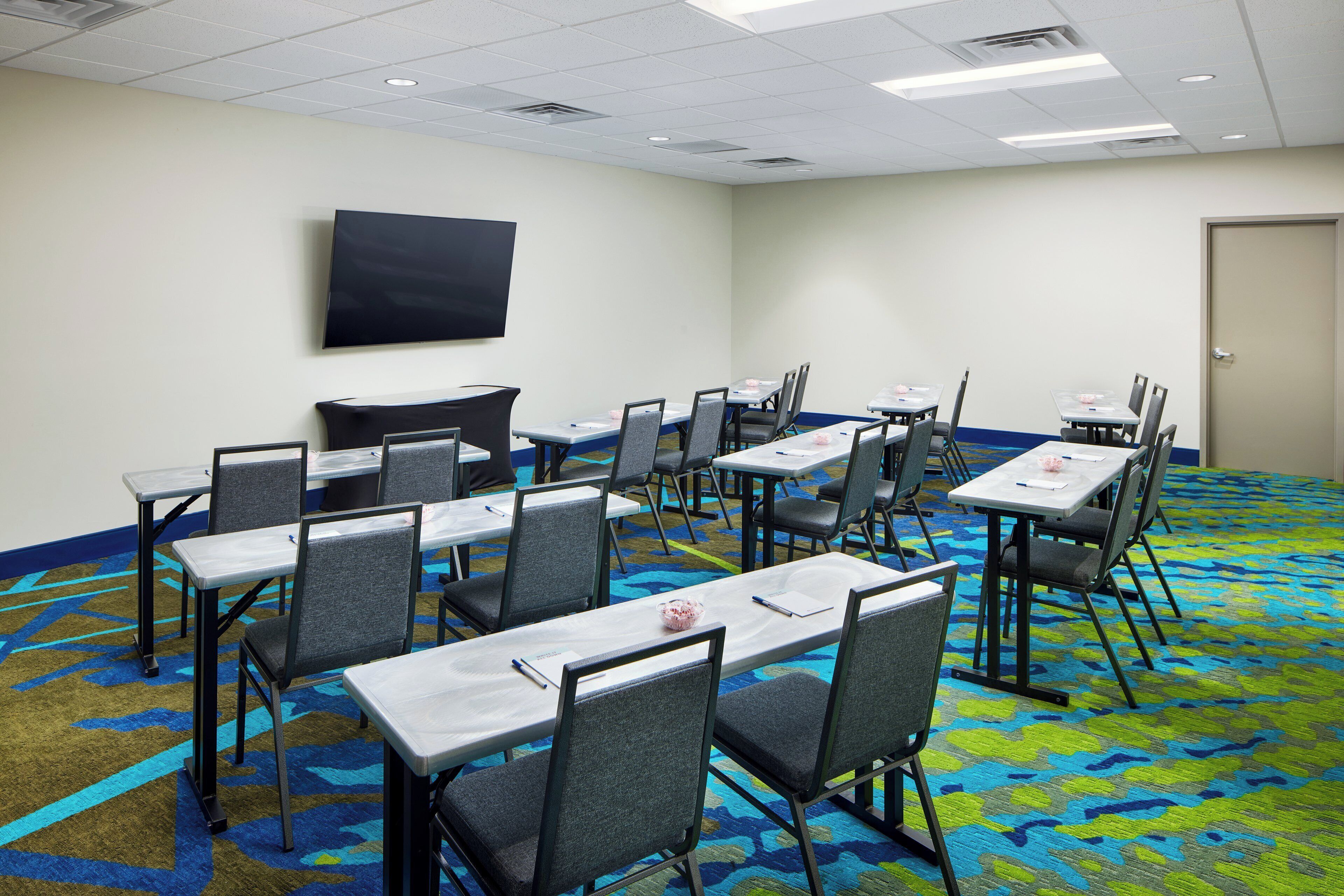 A conference room with tables and chairs, a flat-screen TV on the wall, and a patterned carpet.