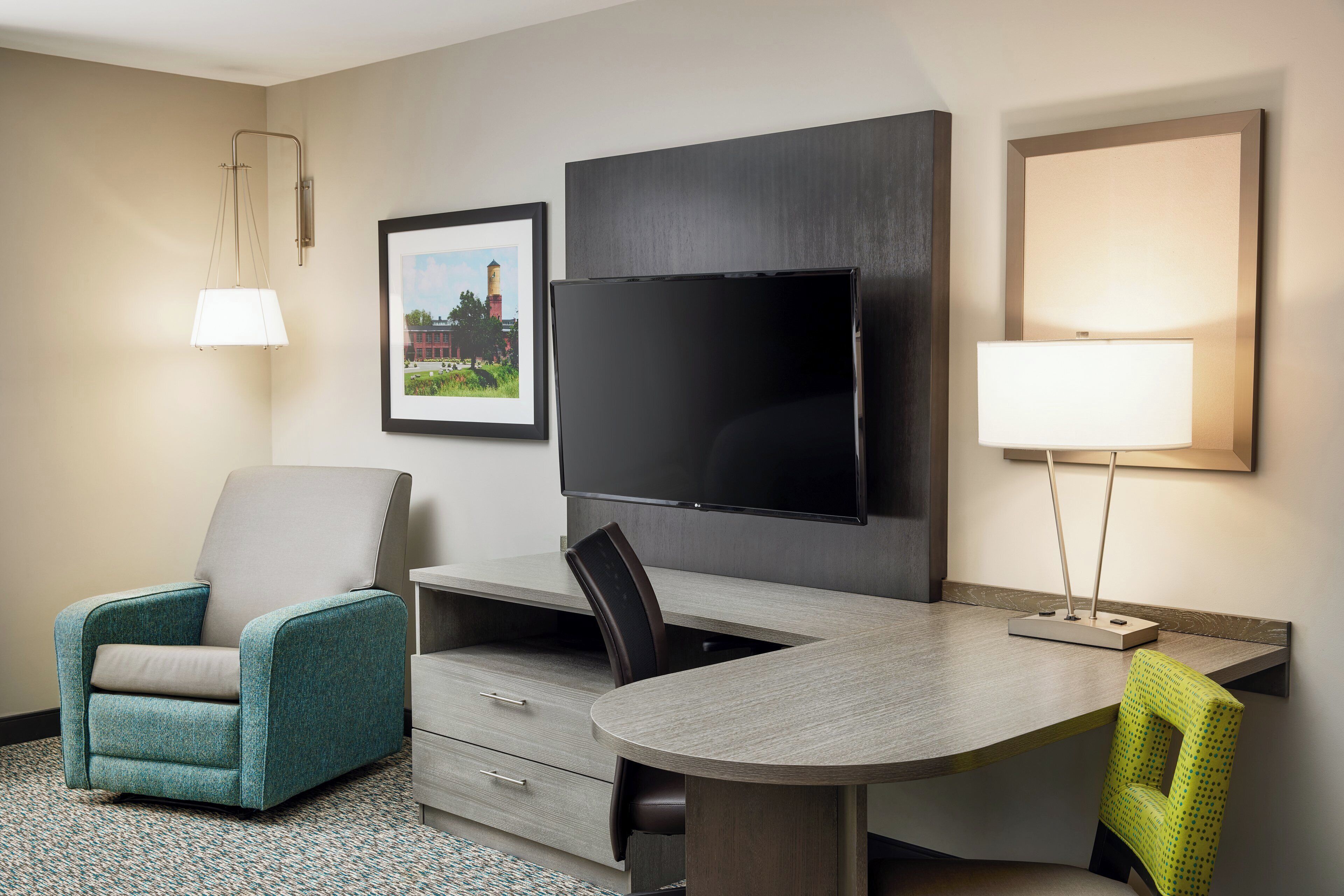 Et hotelværelse med et fladskærms-tv ophængt på væggen, et skrivebord med stol, en turkis lænestol og en lampe.