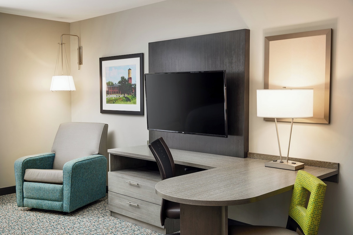 Habitación de hotel con televisor de pantalla plana en la pared, un escritorio con silla, un sillón verde azulado y una lámpara.