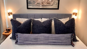 Cama bem arrumada com almofadas escuras e manta listrada, quadro com vista urbana emoldurada acima, e luminárias de leitura fixadas nas paredes em ambos os lados.