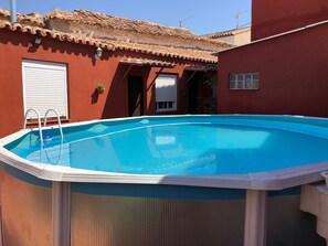 Piscina com um deck branco, uma escada e um prédio vermelho com telhado de azulejos ao fundo.