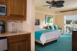Habitación de hotel con kitchenette, cama con cubrecama a rayas, balcón con vistas y ventilador de techo.