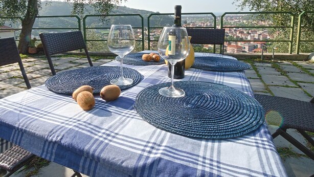 Een tafel buiten gedekt met een geruit tafelkleed, twee wijnglazen, een fles en brood op een terras met uitzicht.