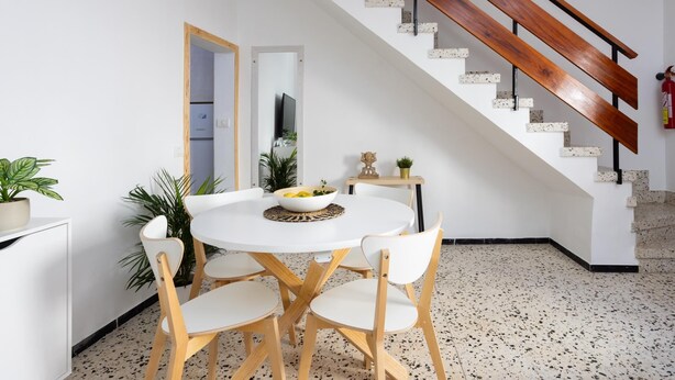 Un área de comedor con una mesa redonda y cuatro sillas, una escalera con escalones de madera, y un extintor en la pared.