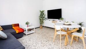 Una sala moderna con un sofá gris, una mesa de comedor blanca con cuatro sillas, un televisor de pantalla plana en la pared y un mueble blanco pequeño con una planta.