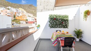 Un balcón con una mesa lista para el desayuno, vistas a un pueblo en la ladera y una pared verde con plantas.