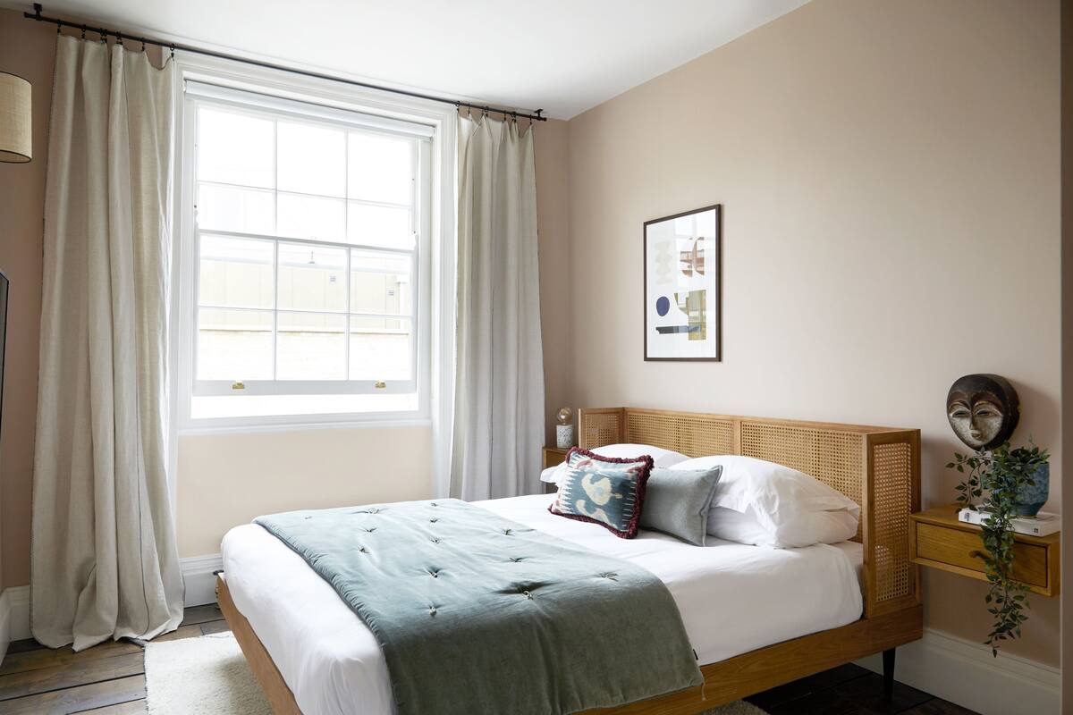 A bedroom with a bed, curtains, a bedside table, a lamp, and a wall-mounted picture.