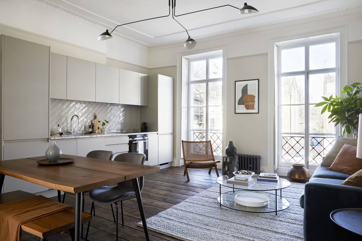 A modern kitchen with white cabinets, a dining area with a wooden table and chairs, a living room with a blue sofa and a coffee table, and large windows with a view of the outside.