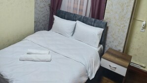 A neatly made bed with white linens and a folded towel, a wooden nightstand with a drawer, and a window with purple and white curtains. - Lorin Otel (Istanbul)