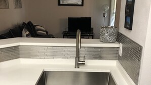 A kitchen with a white countertop, stainless steel sink, and a tiled backsplash. There is a flat-screen TV on a stand in the background.