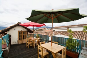 Un patio en la azotea con mobiliario de madera, una sombrilla roja y verde, y una planta en maceta.