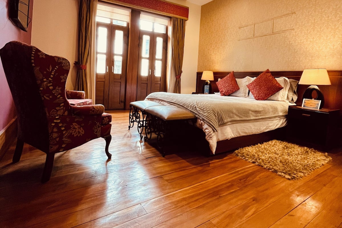 A bedroom with a wooden floor, a bed with red and white bedding, a chair, a nightstand with a lamp, and a window with curtains.