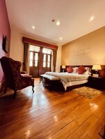 Un dormitorio con suelo de madera, cama con ropa de cama roja y blanca, una silla, una mesita de noche con lámpara y una ventana con cortinas.