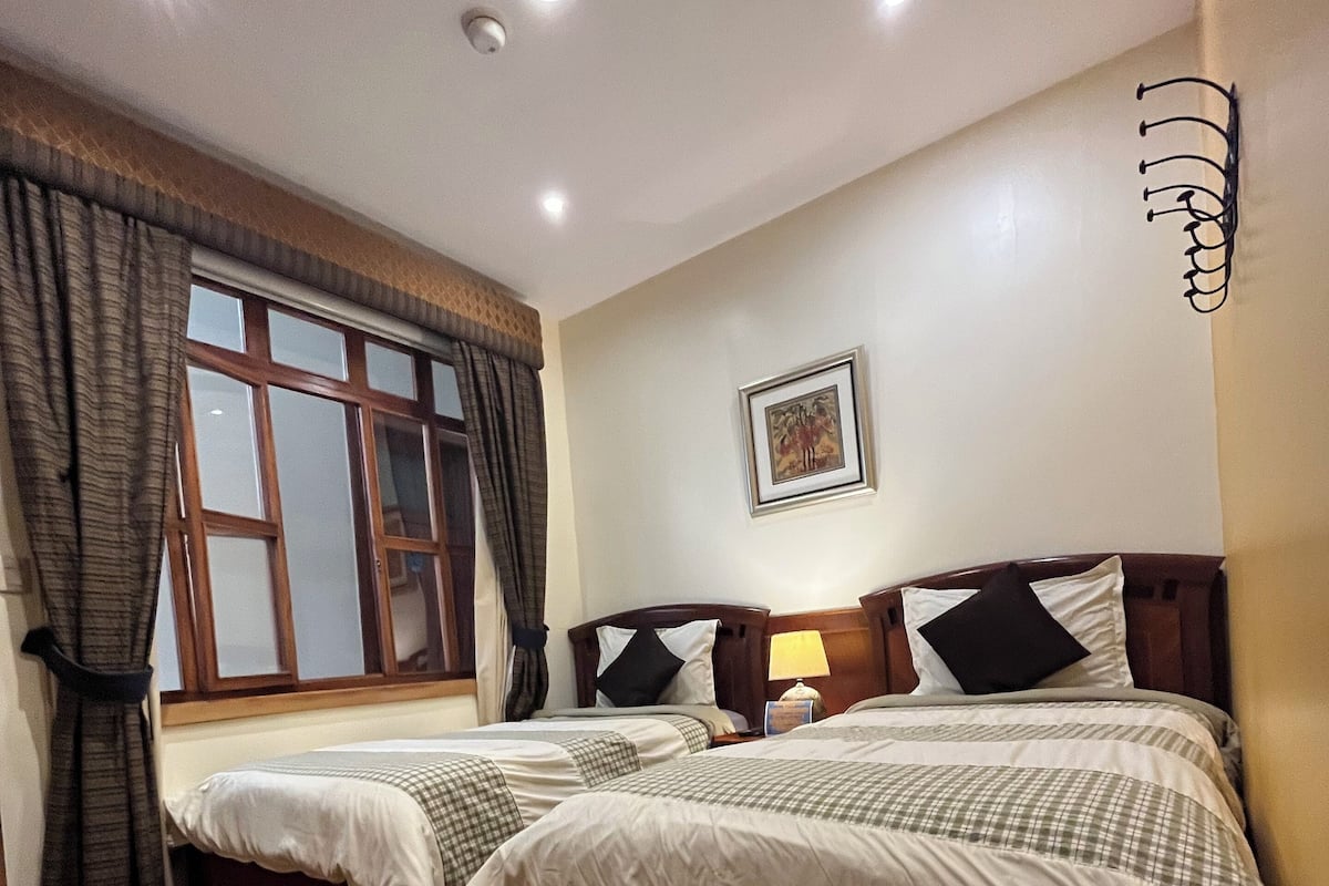 A hotel room with two wooden beds, a window with curtains, a framed picture on the wall, and a ceiling light.