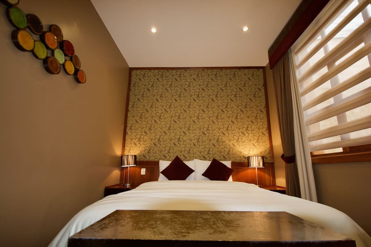 A hotel room with a bed, two bedside lamps, a wall with a patterned wallpaper, and a window with blinds.