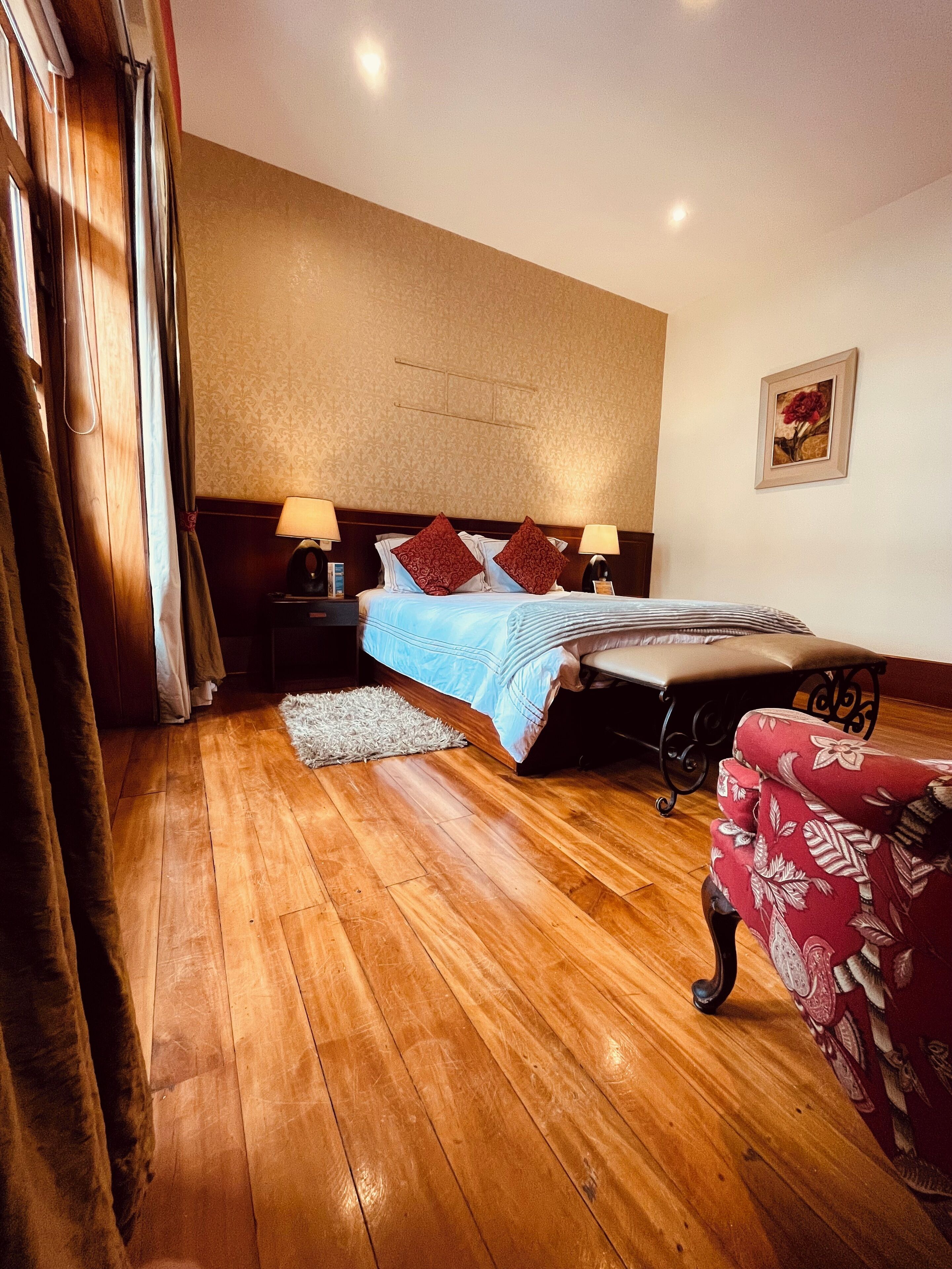 Une chambre avec un plancher en bois, un lit recouvert d’une couvre-lit bleue, deux oreillers rouges, une table de chevet avec une lampe, un fauteuil rouge recouvert de tissu et un tableau encadré accroché au mur.