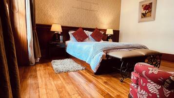 Une chambre avec un plancher en bois, un lit recouvert d’une couvre-lit bleue, deux oreillers rouges, une table de chevet avec une lampe, un fauteuil rouge recouvert de tissu et un tableau encadré accroché au mur.