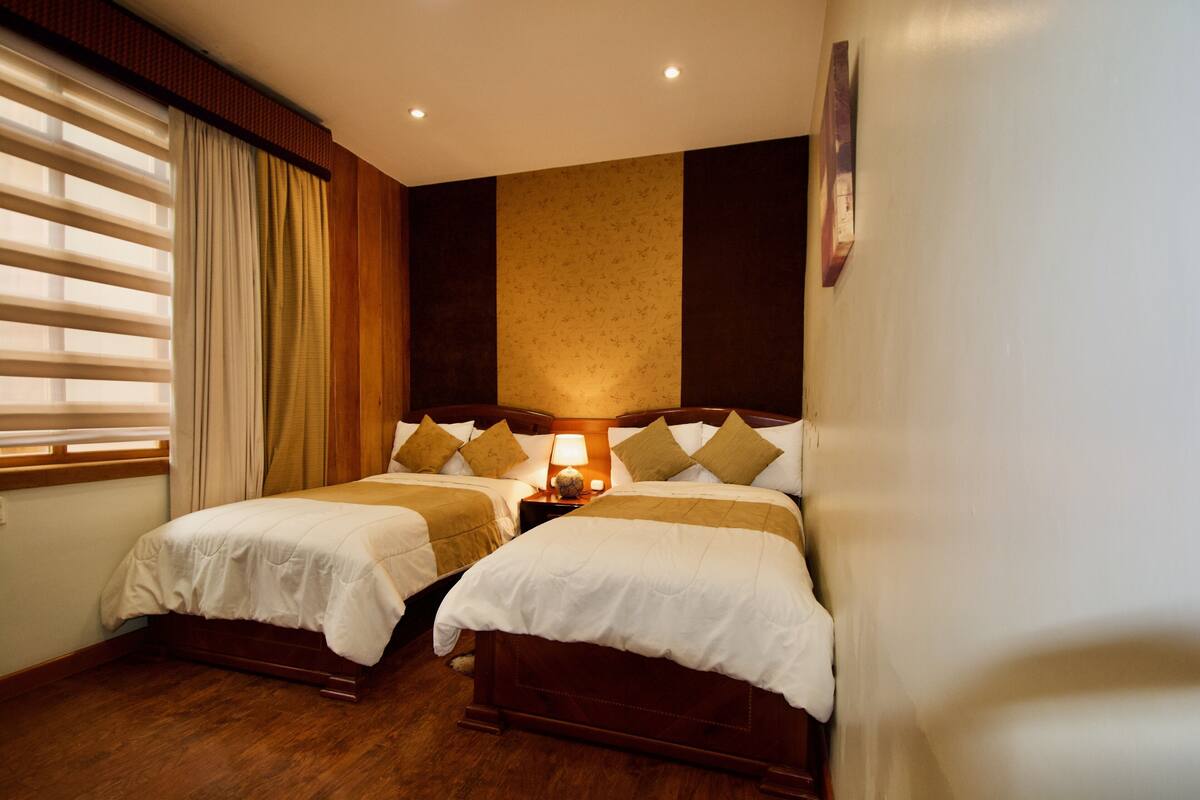 A double room with two beds, a nightstand with a lamp, wooden flooring, and a window with blinds.