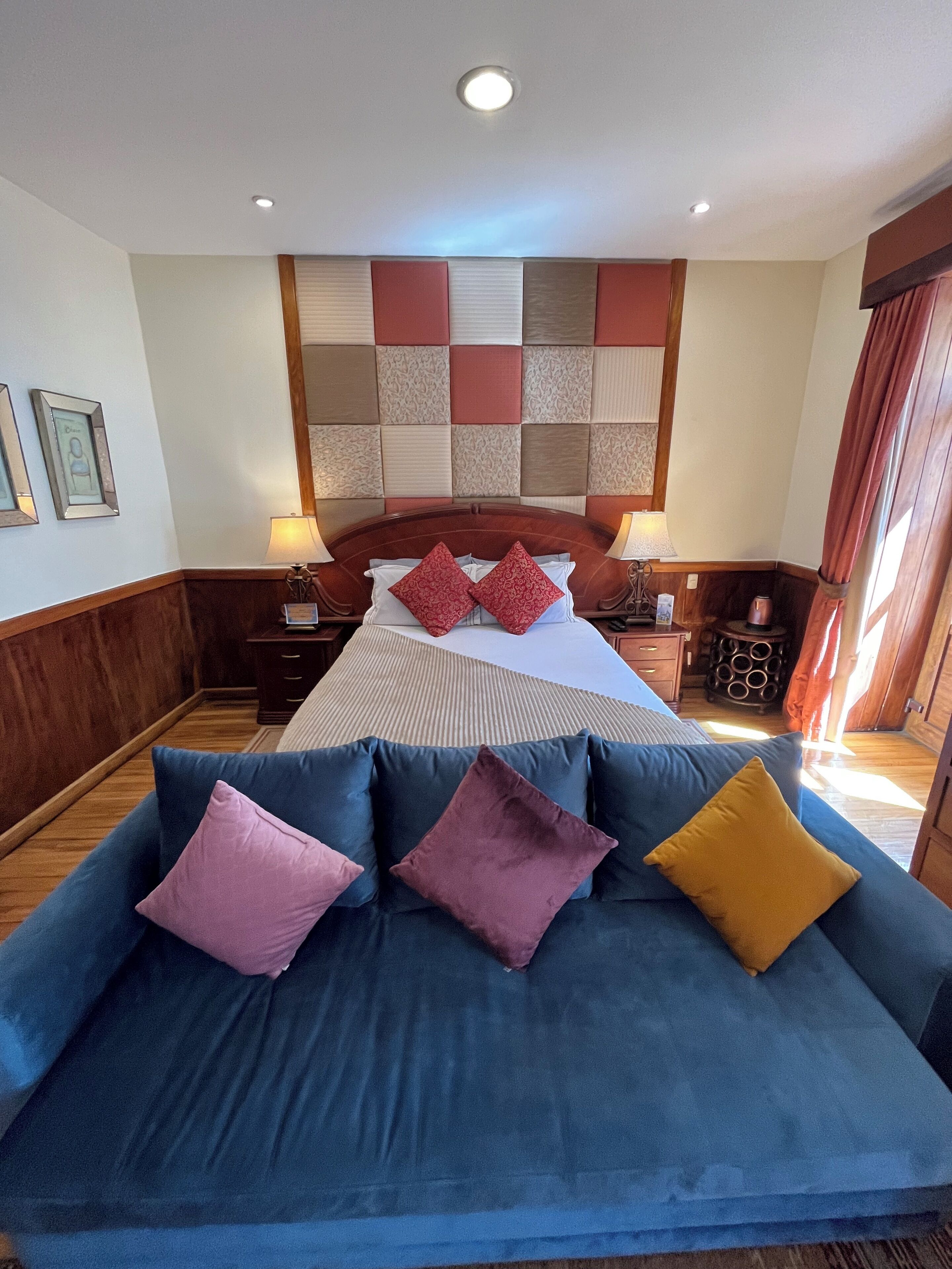 A hotel room with a large bed, a blue sofa with multiple cushions, a wooden headboard, and a decorative wall panel.