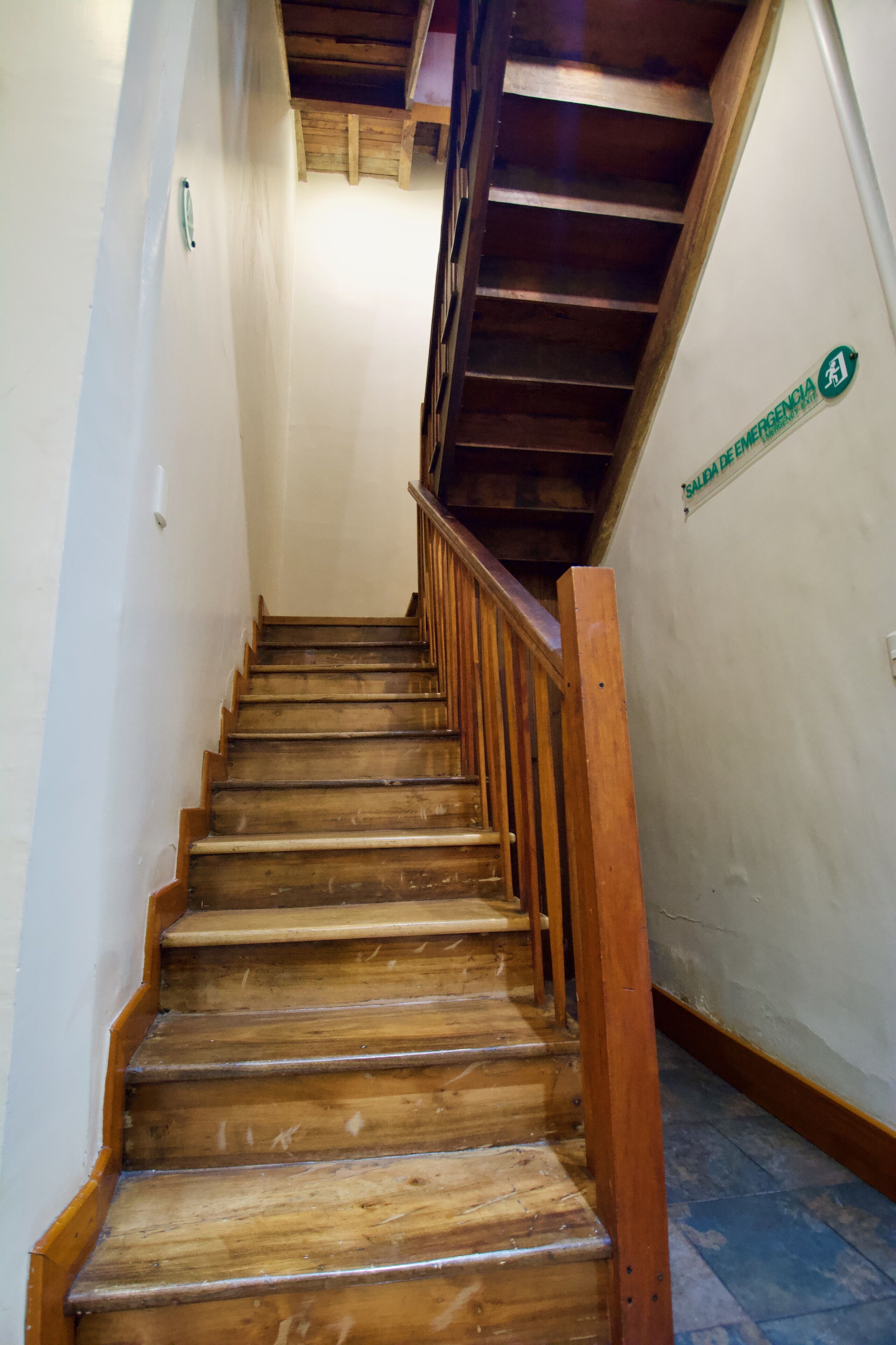 Un escalier en bois muni d’une rampe, menant à un palier où se trouve une signalisation indiquant une sortie de secours.