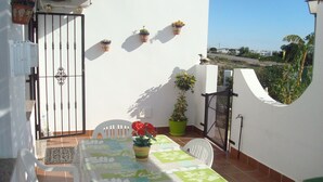 Un balcon avec une table dressée pour deux personnes, entouré de chaises blanches, d’une plante en pot et offrant une vue sur la ville.