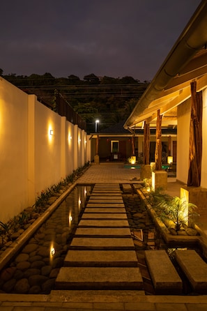 Un chemin illuminé la nuit qui mène à un bâtiment doté d’une piscine, entouré de murs et de plantes.