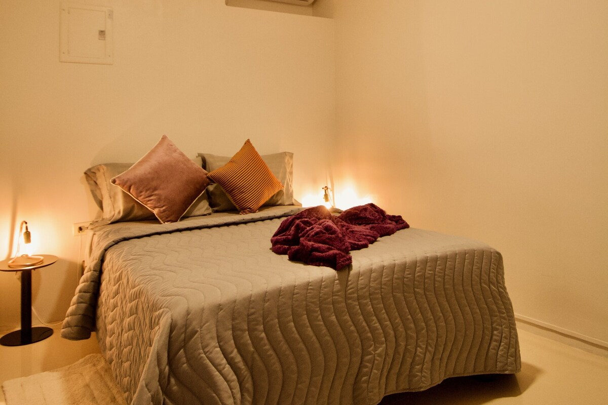 A neatly arranged bedroom with a bed, pillows, a bedside table, and a wall-mounted air conditioner.