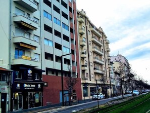 Un immeuble à plusieurs étages doté de balcons, un magasin avec une enseigne indiquant « Optic 2000 », et une rue bordée de voitures et d’un espace vert.