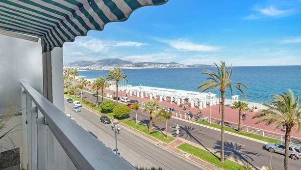 Un balcon donnant sur une promenade en bord de mer, avec des palmiers et un ciel bleu dégagé.