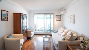 Un salon spacieux doté d’un plancher en bois, d’un canapé blanc, d’un fauteuil blanc et d’une porte en verre donnant sur un espace extérieur.