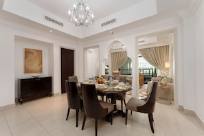 Un coin repas avec une table ronde dressée pour six personnes, entourée de chaises sombres, d’un lustre et d’un grand tableau abstrait accroché au mur.