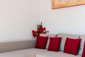 Un rincón de una habitación con un sofá color beige y cojines rojos, un jarrón pequeño con flores rojas, y un cuadro en la pared.