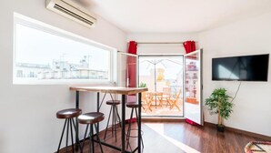 Une pièce avec une table en bois et des tabourets de bar, un téléviseur fixé au mur et une vue sur un balcon extérieur agrémenté de rideaux rouges.