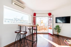 Une pièce avec une table en bois et des tabourets de bar, un téléviseur fixé au mur et une vue sur un balcon extérieur agrémenté de rideaux rouges.
