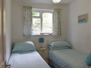 A bedroom with two single beds, a window with floral curtains, a bedside lamp, and a painting on the wall.