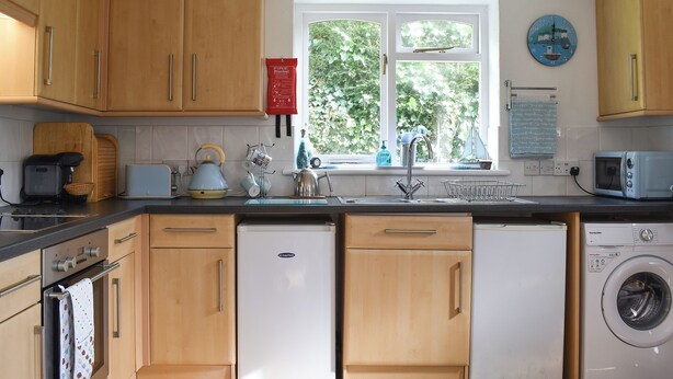 A kitchen with wooden cabinets, a washing machine, a microwave, and a window with a view of greenery. - 2 bedroom accommodation in Talland Bay, near West Looe (Talland Bay, near West Looe)