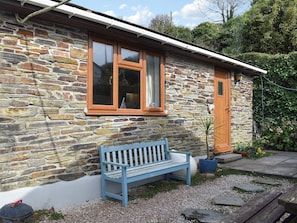 A stone house with a wooden bench in front, a door, and a window with a curtain.