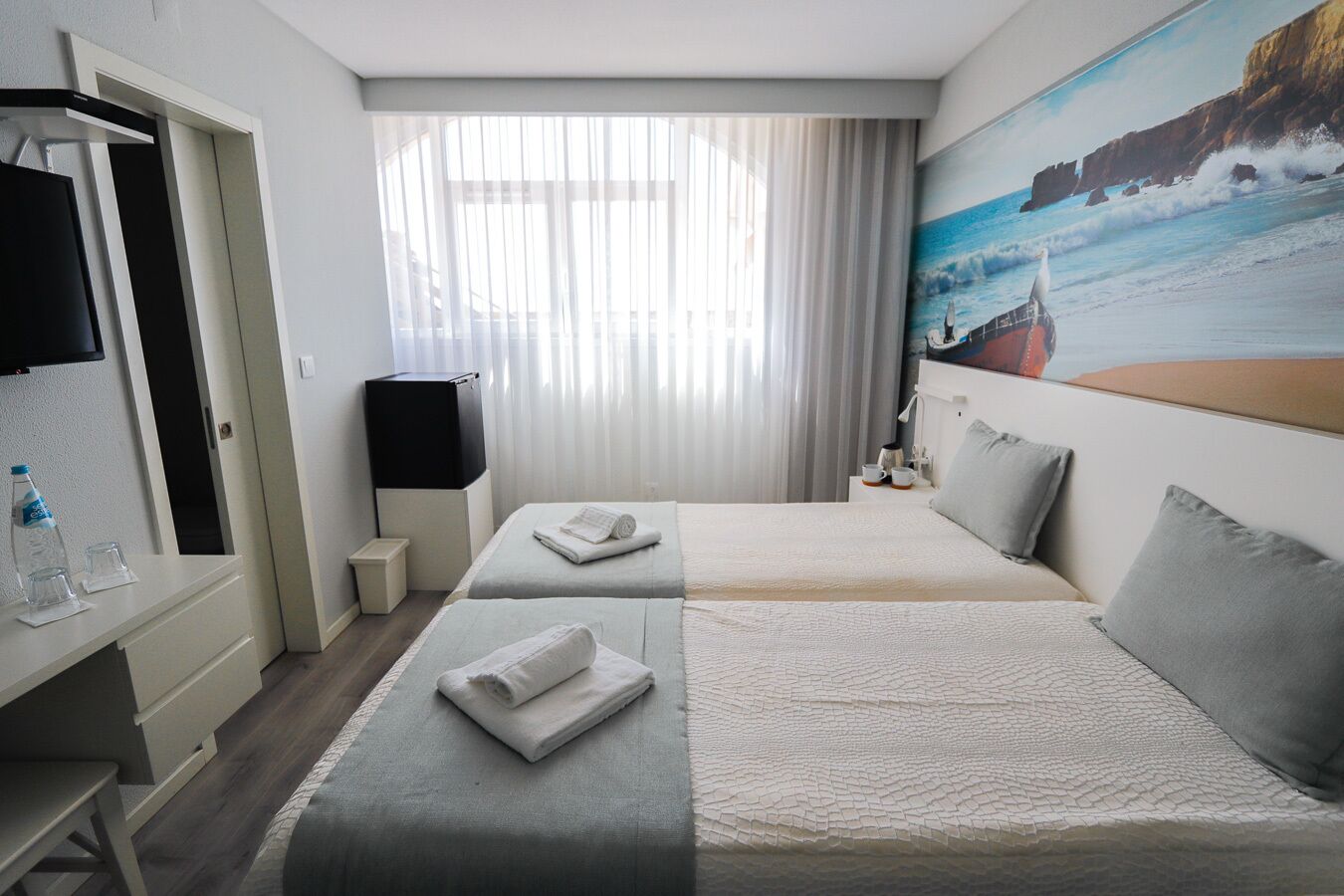 Hotellihuone, jossa on kaksi sänkyä, televisio, merimaiseman kuvaava taulu ja ikkuna, jossa on läpikuultavat verhot.