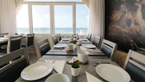 Uma área de refeições com vista para o oceano, com uma mesa comprida posta com pratos, xícaras e um vaso de planta.