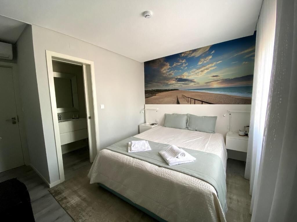 A hotel room with a large bed, a painting of a beach scene, a nightstand, and a door leading to another room.