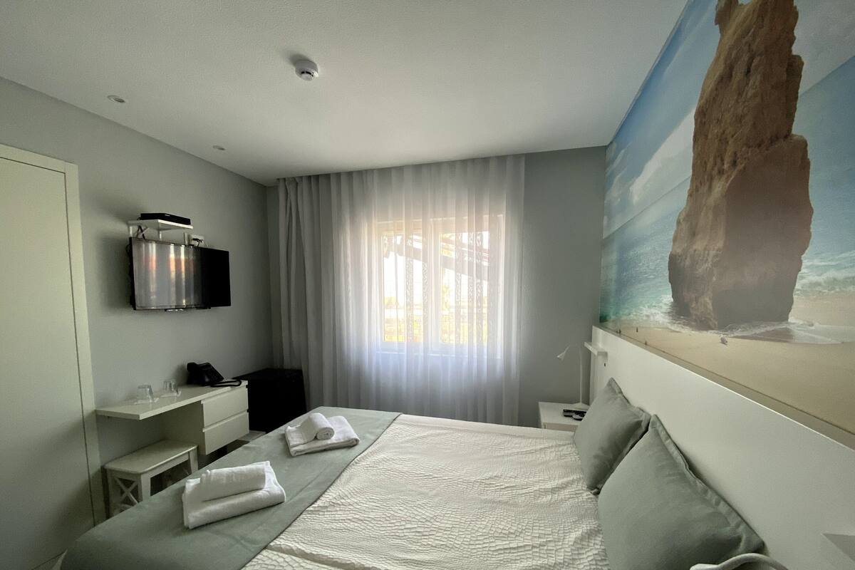 A hotel room with a bed, a flat-screen TV, a desk, a chair, and a painting of a beach scene.