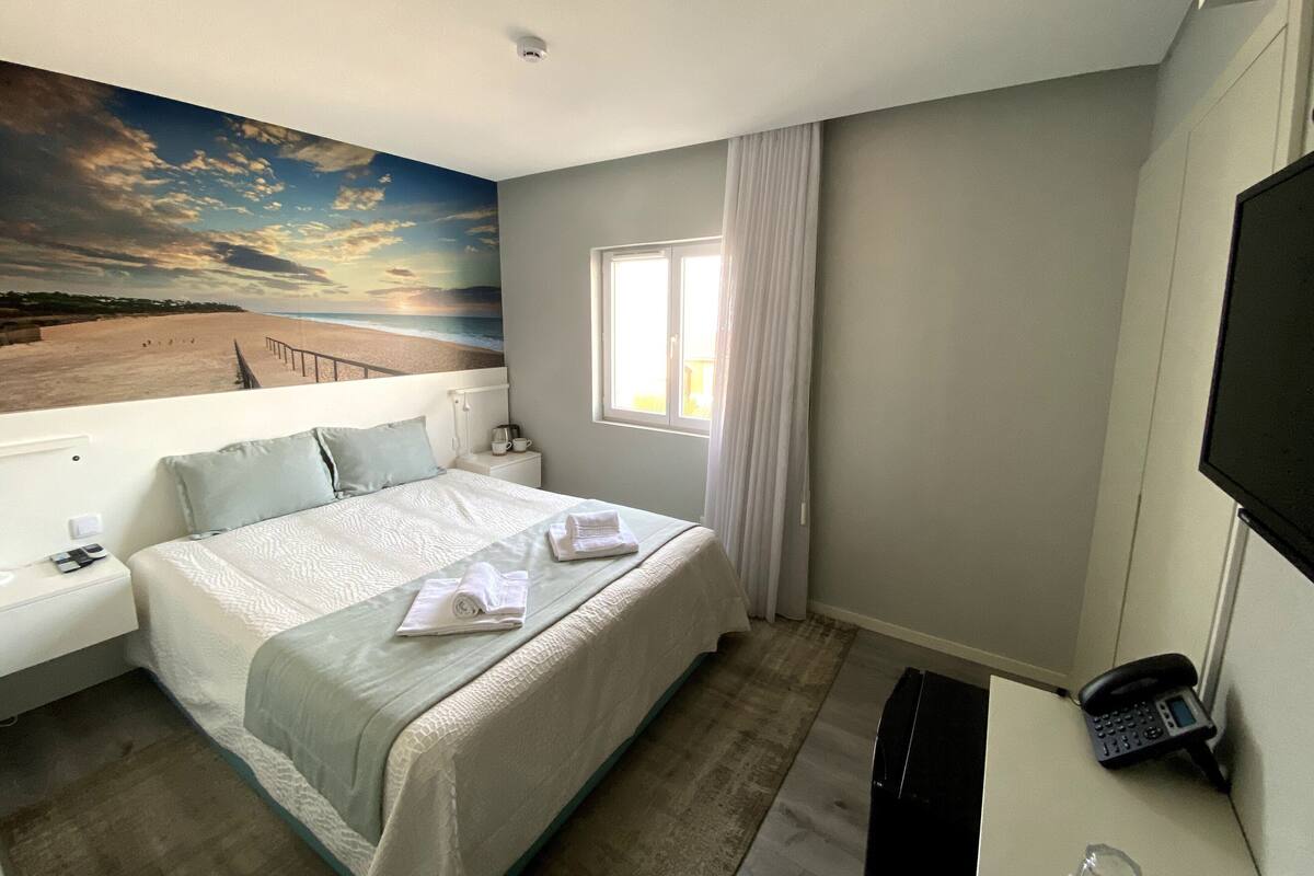 A hotel room with a bed, a nightstand, a telephone, a window with curtains, and a wall mural of a beach scene.