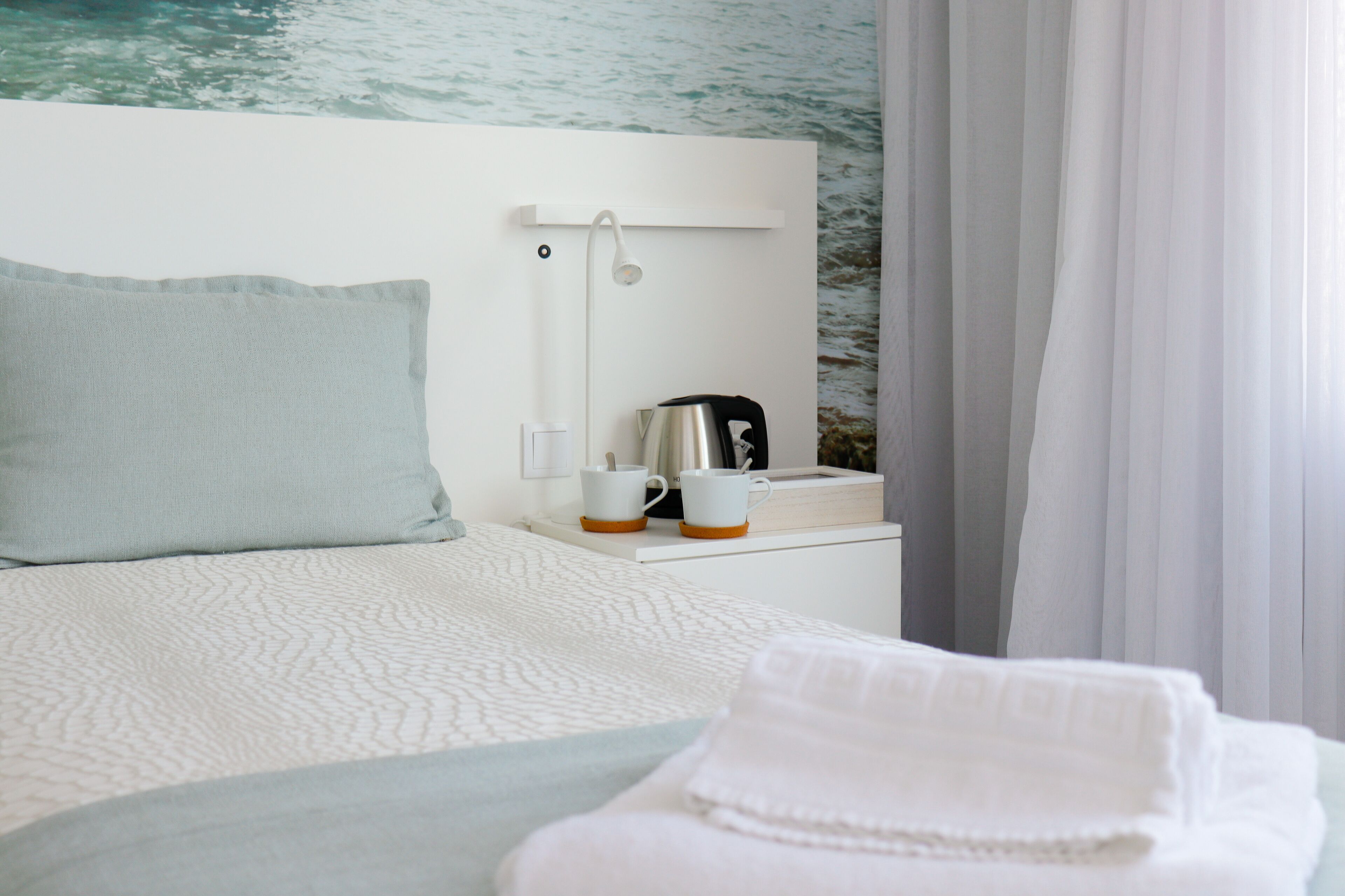 A neatly made bed with a pillow, a bedside table with a kettle, cups, and a small book.