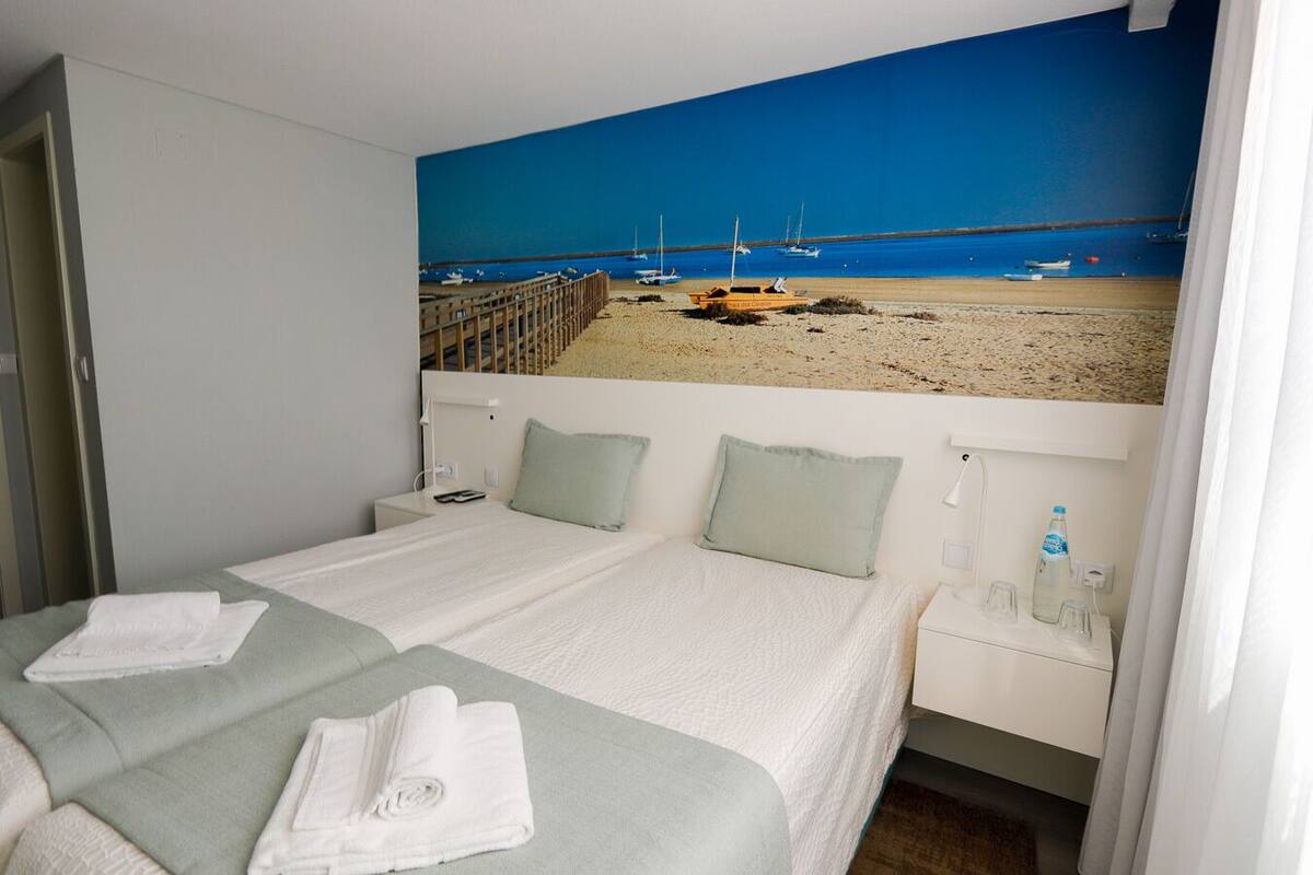 A hotel room with a bed, two pillows, a nightstand, a lamp, and a bottle of water.