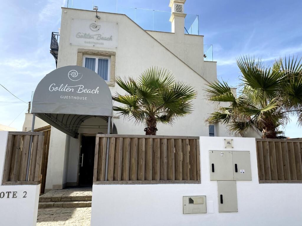 Una casa de huéspedes llamada Golden Beach con fachada blanca, palmeras y una valla de madera.