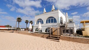 Um edifício à beira-mar com uma escadaria que leva à entrada, palmeiras e guarda-sóis de praia.